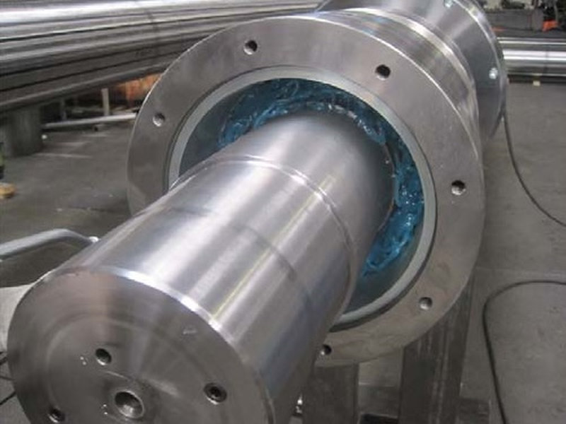 Applicazione di lubrificante, olio industriale ad alta viscosità su un albero meccanico. | Svecom Primo piano di un grosso albero meccanico inserito in un cilindro flangiato con olio industriale e grasso lubrificante blu all'interno.